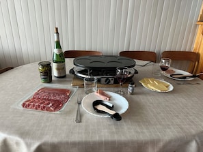 A dining table set with a bottle of wine, a jar of olives, a plate of sliced meat, a cheese board, and a portable grill.