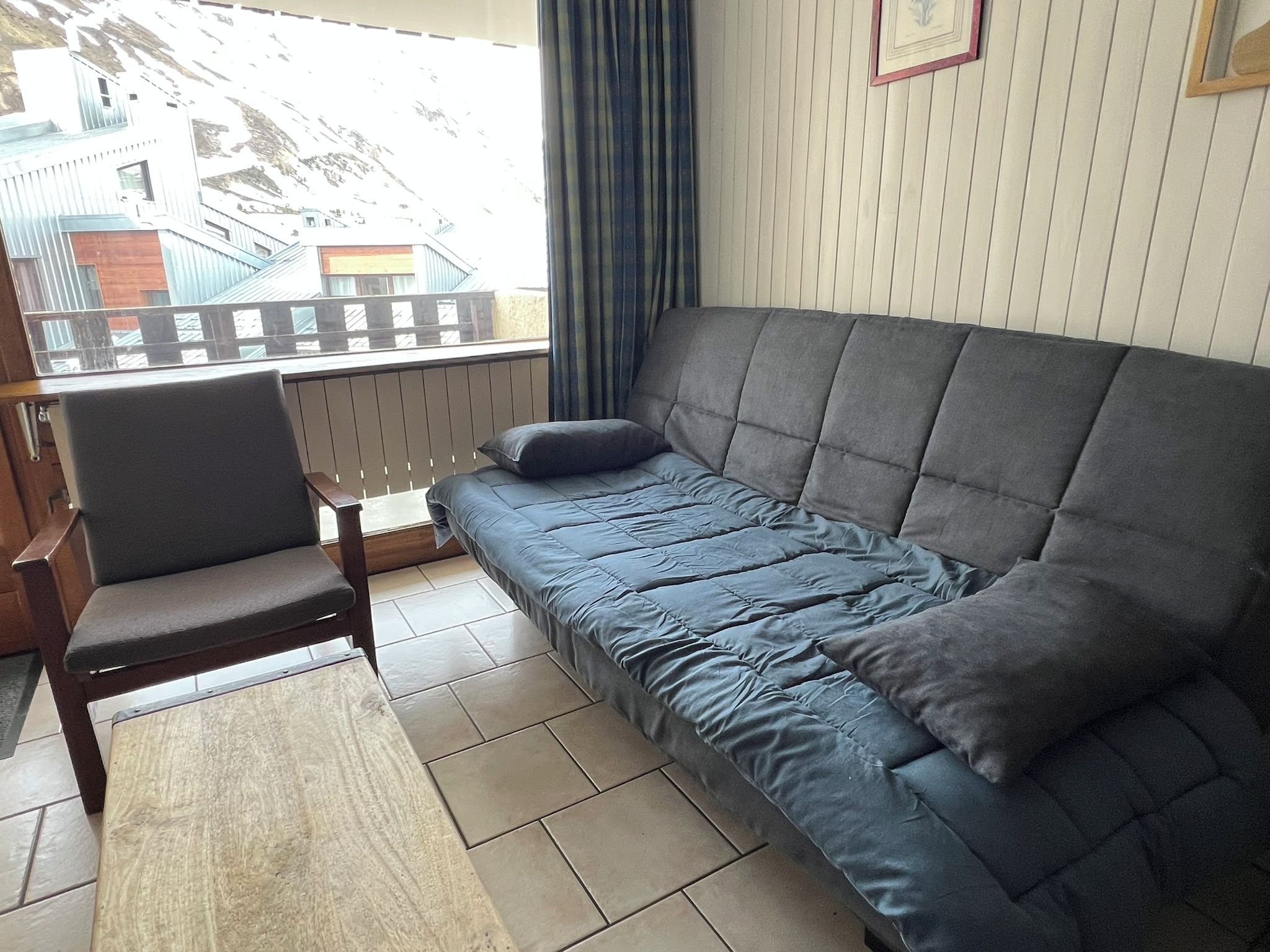Een kamer met een bank, een stoel, een raam met uitzicht op gebouwen en een gordijn.