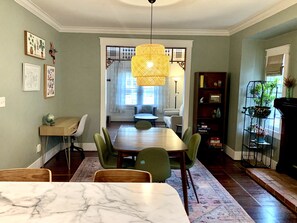 A dining area with a marble table, green chairs, a hanging rattan light fixture, and a bookshelf with plants.