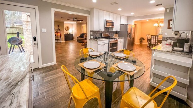 A modern kitchen with white cabinets, a stainless steel refrigerator, and a dining area with a glass table and yellow chairs. - Retreat in the woods! (Houston)