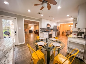 A modern kitchen with white cabinets, a stainless steel refrigerator, and a dining area with a glass table and yellow chairs. - Retreat in the woods! (Houston)