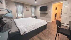 A bedroom with a bed, a desk with a lamp, a chair, a ceiling fan, a TV, and a door leading to another room. - Retreat in the woods! (Houston)