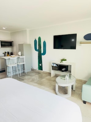 Quarto de hotel moderno com uma cama, pequena área de cozinha com micro-ondas, um banquinho de bar, uma decoração de cacto, uma TV e uma mesa de centro redonda.