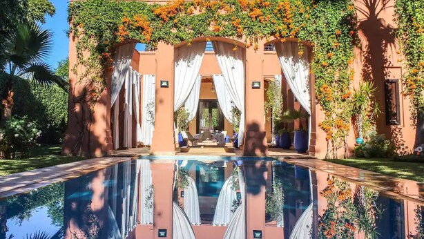 Ein Poolbereich mit spiegelglatter Wasseroberfläche, eine überdachte Lounge mit weißen Vorhängen und üppiges Grün.