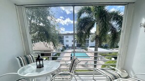 A balcony with a glass table, striped chairs, and a view of a pool and buildings.