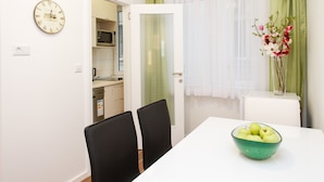 A dining area with a table, chairs, a bowl of apples, a clock, and a kitchen in the background. - Apartment Jovanova (Belgrade)