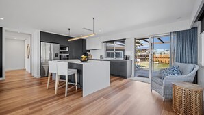 Una cocina moderna con una isla central, taburetes blancos tipo bar y una mesa de comedor. Hay un amplio ventanal corredero que da a un patio trasero.