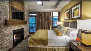 A hotel room with a stone fireplace, a large bed with a gold accent, a nightstand with a lamp, and a view of the outdoors. - Stay and Ski in Northstar Resort (Truckee)