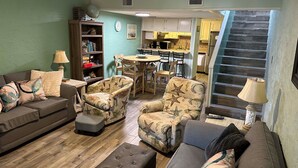 A living room with a sofa, armchairs, a coffee table, and a bookshelf. There is a kitchen in the background with a dining area. - Family&Pre-Approved Pet Friendly Condo Gated Complex Bed 3 Bath 3 walk2Tootsies (Panama City Beach)