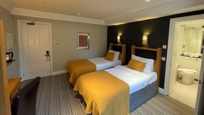 A hotel room with two beds, a desk, a chair, a door, and a bathroom visible through an open door. - Shandon House (Richmond)