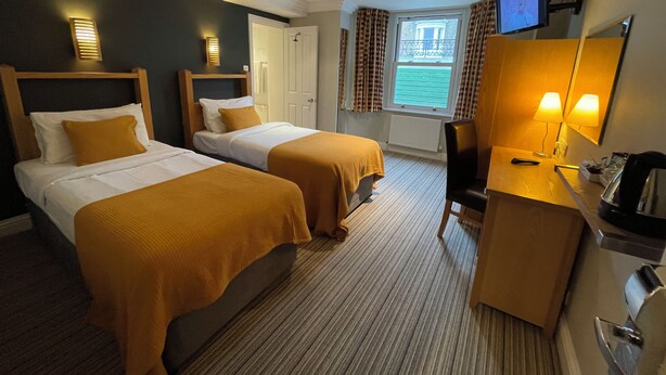 A hotel room with two beds, a desk with a chair, a TV, and a window with curtains. - Shandon House (Richmond)