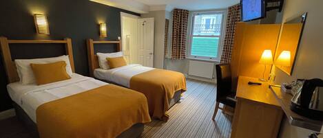 Quarto de hotel com duas camas, uma mesa de trabalho com cadeira, televisão e janela com cortinas.