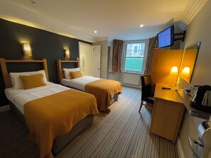 A hotel room with two beds, a desk with a chair, a TV, and a window with curtains. - Shandon House (Richmond)