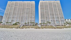 Deux immeubles de grande hauteur dotés de nombreux balcons, une plage de sable au premier plan et des palmiers.