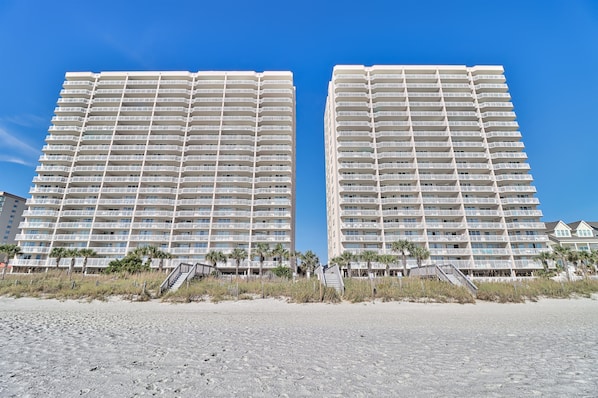 Dos edificios altos con balcones, una playa arenosa al frente y palmeras.