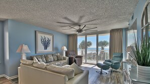 Un salon spacieux doté d’un ventilateur de plafond, d’une grande fenêtre offrant une vue sur l’océan et d’un balcon avec une chaise et un pot de fleurs.