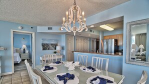 Une salle à manger avec une grande table dressée pour six personnes, un lustre et une cuisine visible à travers une porte ouverte.