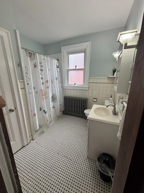 A bathroom with a shower curtain, a window, a radiator, a sink, and a trash bin.