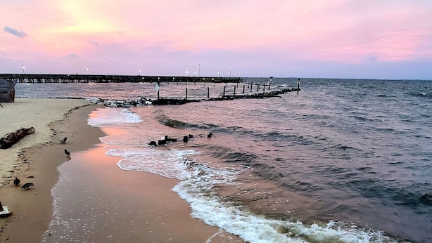 A beach with a wooden pier extending into the water, waves gently lapping at the shore, and a sunset in the background. - Luxury Cottage w/ Bay & Boardwalk Views--steps from the water (North Beach)