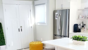 A modern kitchen with a stainless steel refrigerator, a white countertop, and a yellow ottoman. There are two wooden stools at the kitchen island. - Luxury Cottage w/ Bay & Boardwalk Views--steps from the water (North Beach)
