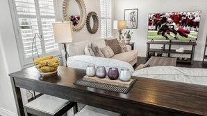 A living room with a dark wooden dining table, cushioned chairs, a sofa, and a television displaying a football game. - Milton Home away from home (Milton)