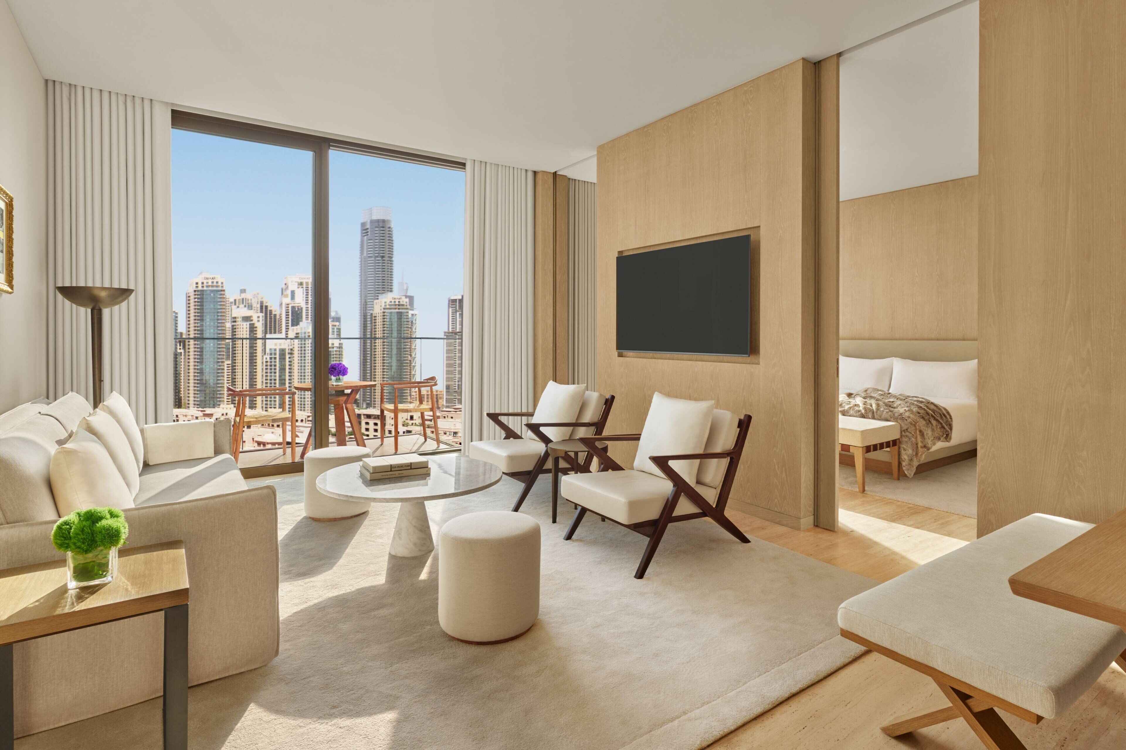 A modern hotel room with a large window offering a city view, a sofa, a dining table, and a bed.