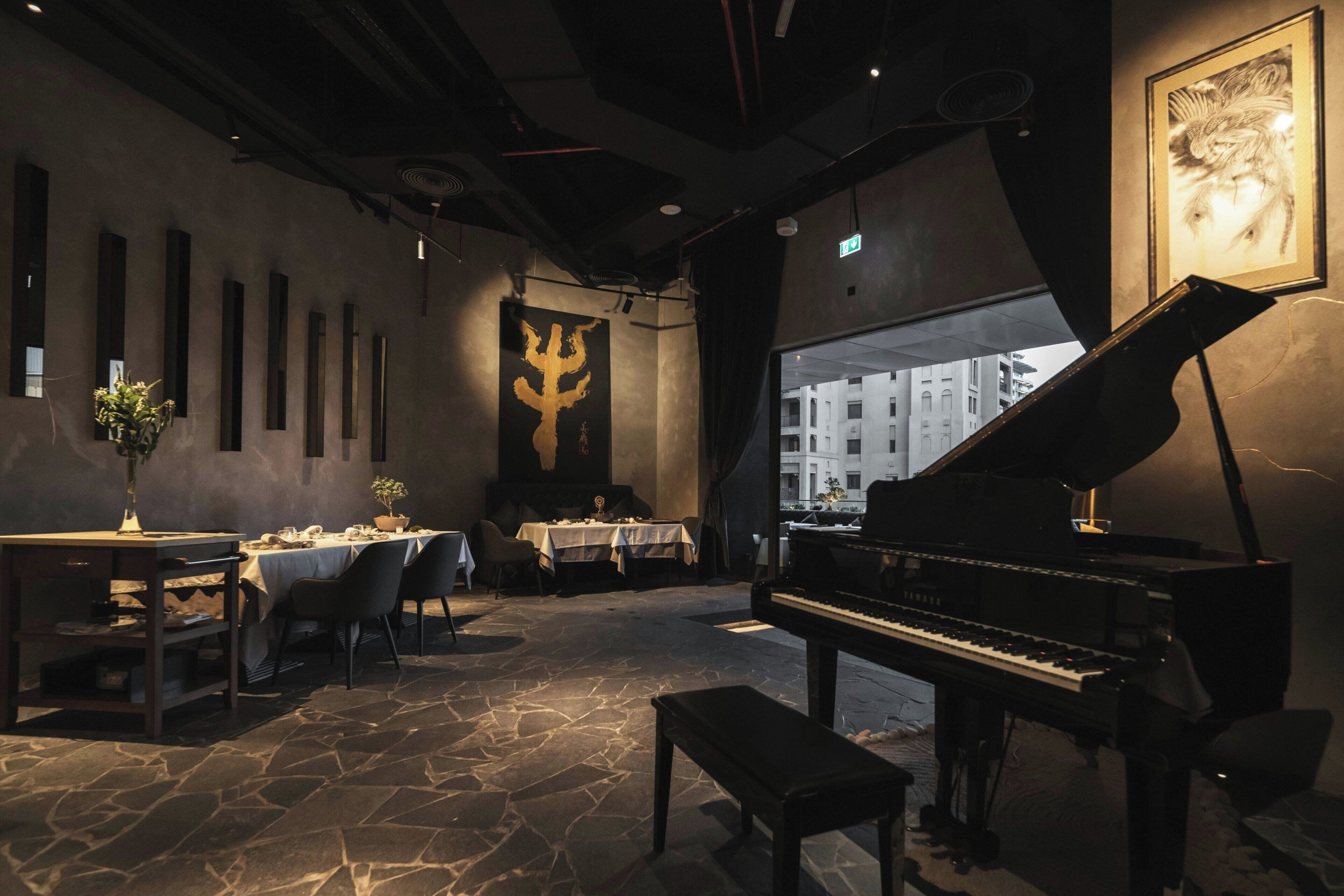 A restaurant with a grand piano, stone flooring, tables with white tablecloths, and a large wall decoration.