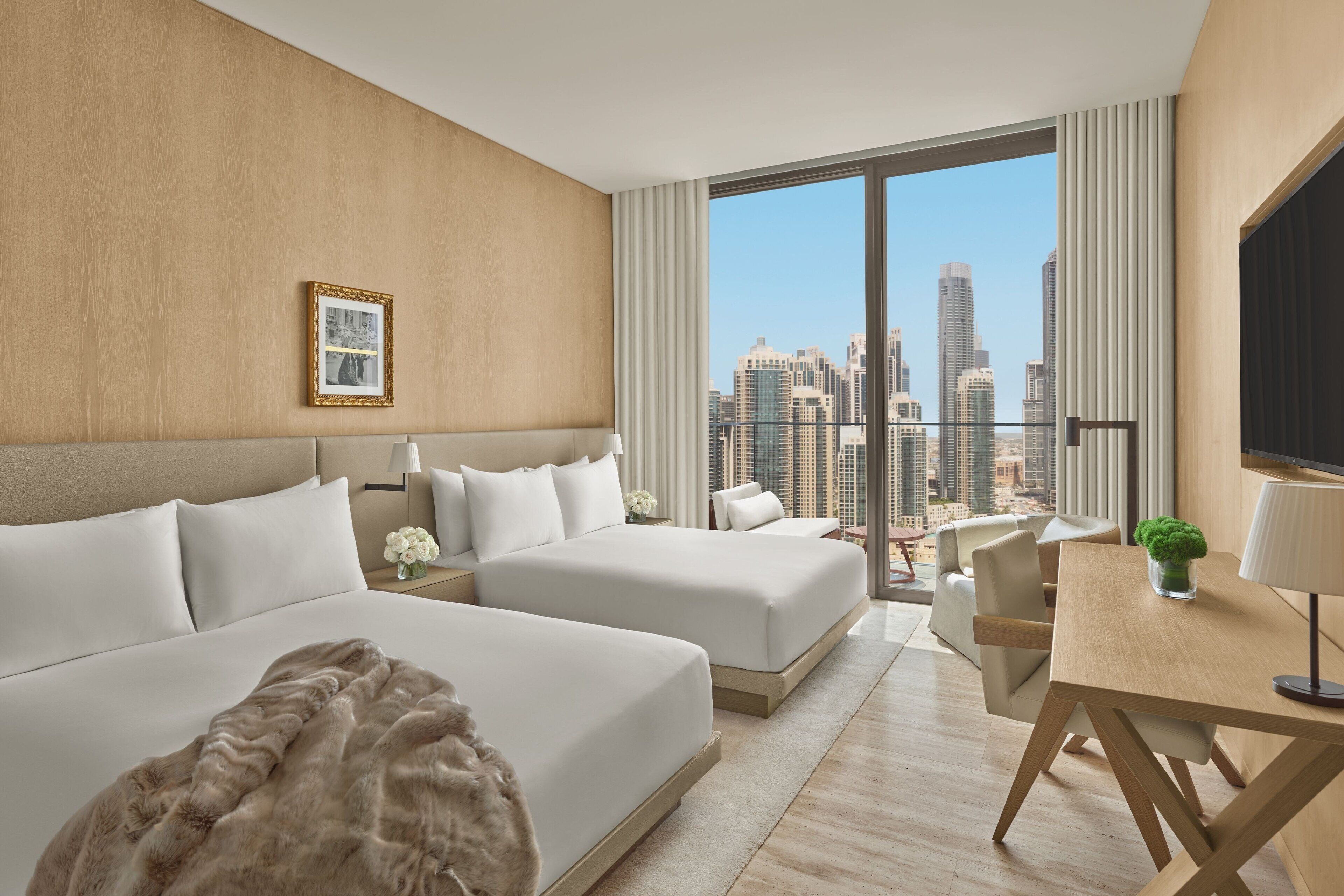 A hotel room with two beds, a desk, a TV, and a view of the cityscape.