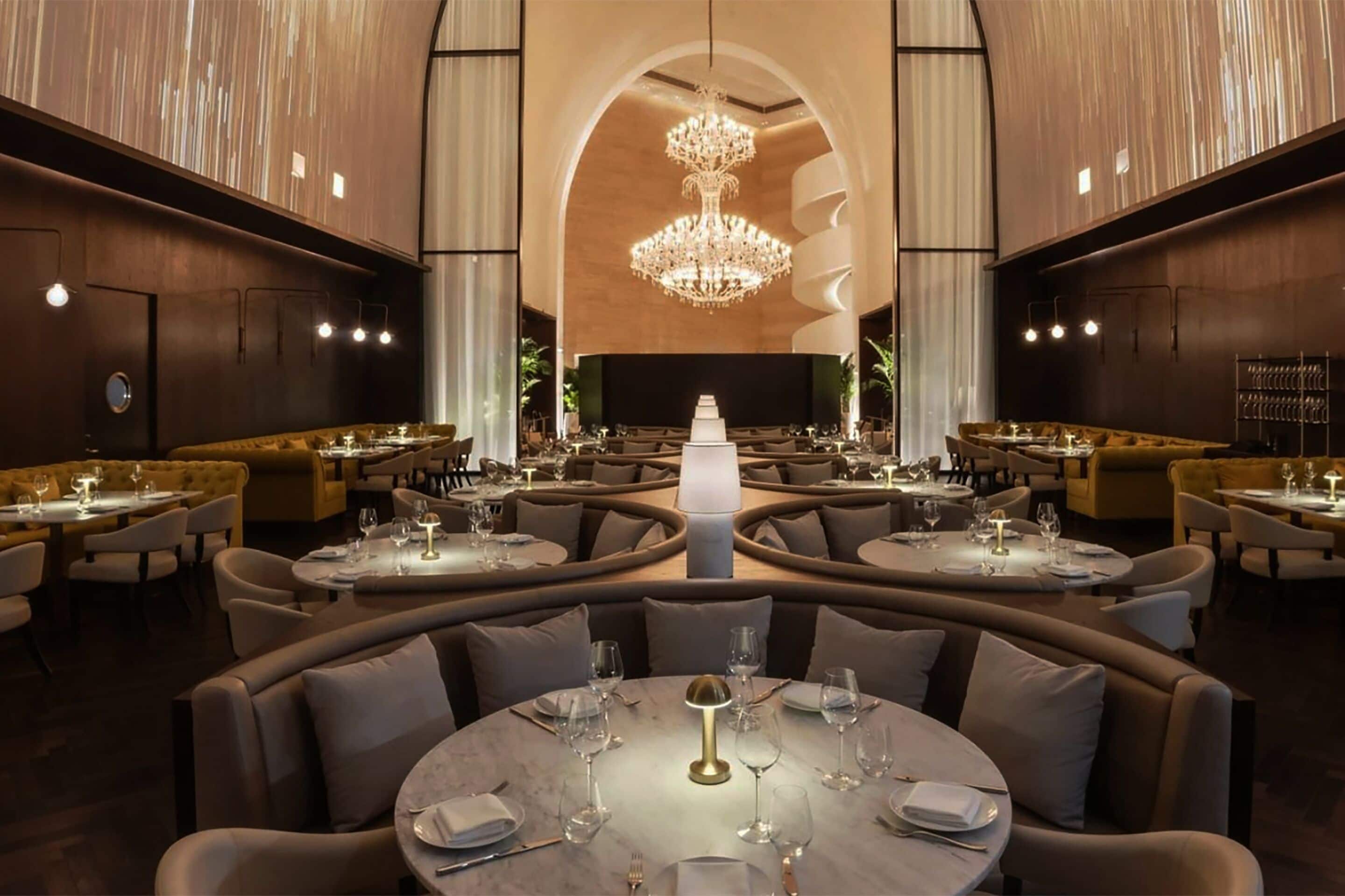 A spacious, elegantly designed restaurant with round tables, comfortable seating, and a central chandelier.
