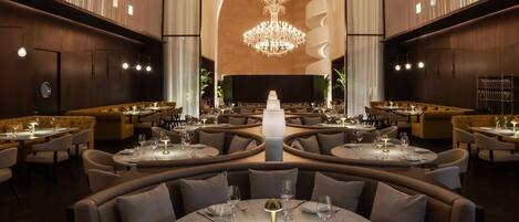 A spacious, elegantly designed restaurant with round tables, comfortable seating, and a central chandelier.