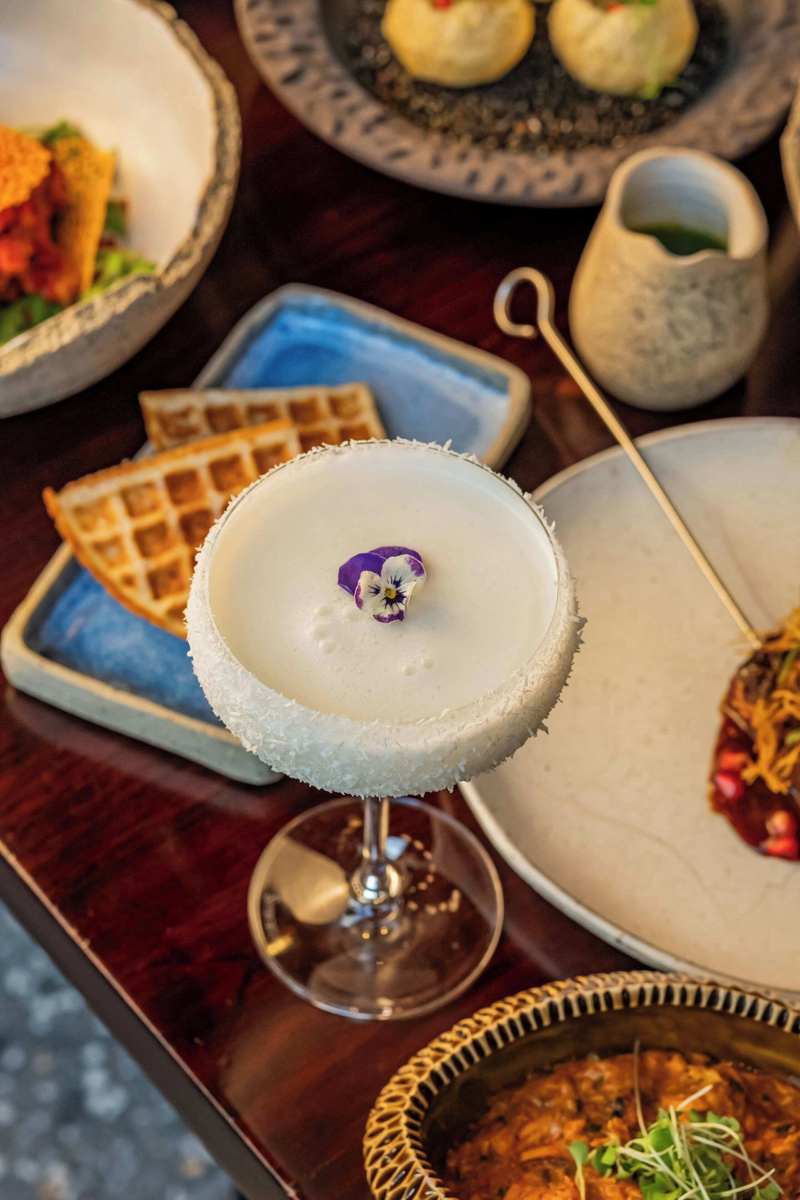 A dessert with a white, frosted top and a purple garnish, accompanied by waffles and a bowl of soup.
