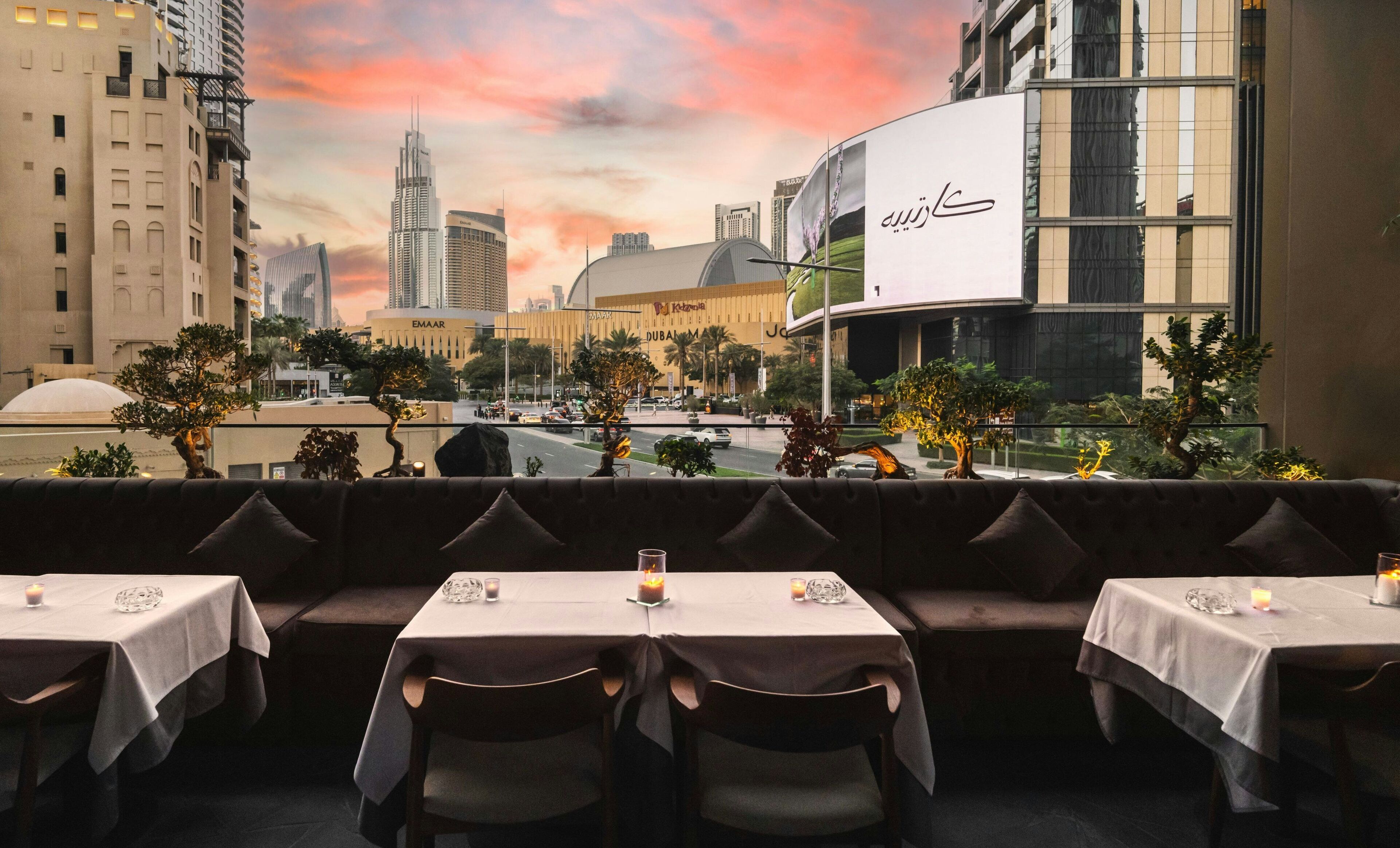 Un restaurante con mesas dispuestas para comer, con vistas a un paisaje urbano de edificios modernos y un cielo al atardecer.