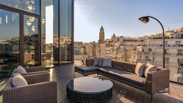 A modern rooftop terrace with a large glass wall, a sectional sofa, a round coffee table, and a street lamp. - Bvlgari Hotel Paris (Paris)