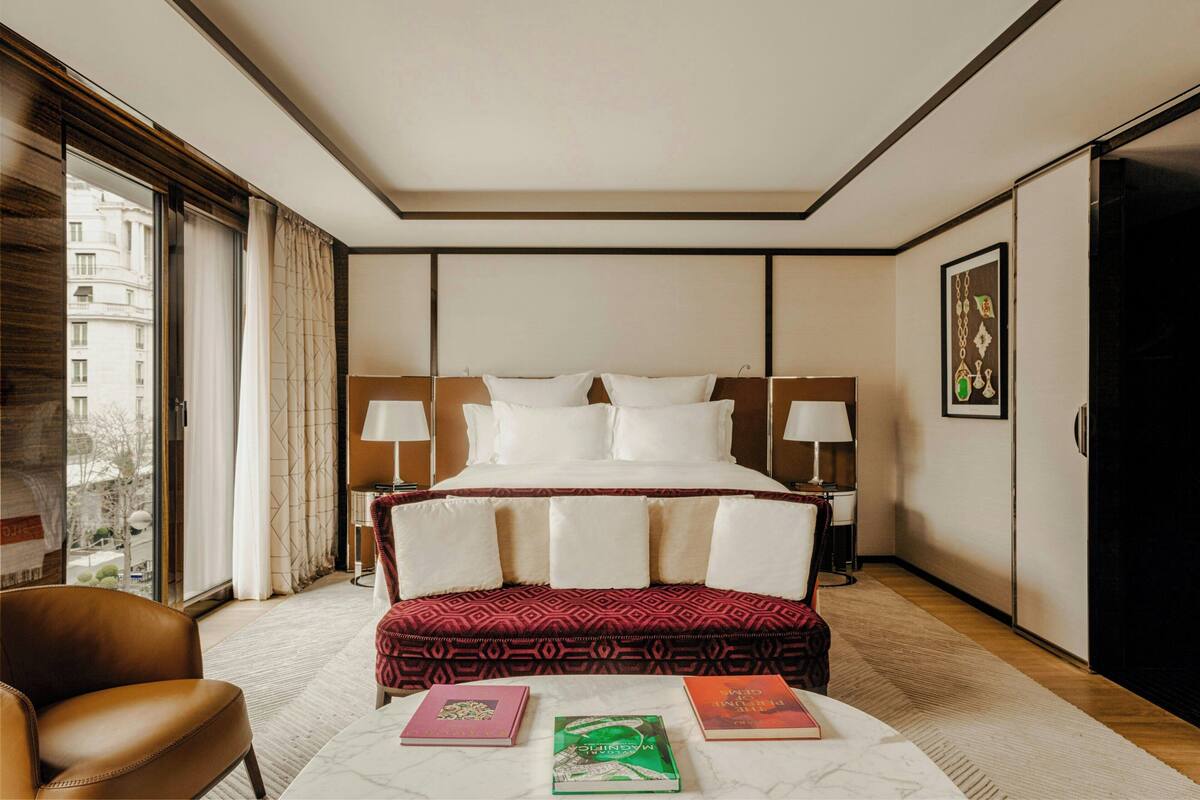 A hotel room with a large bed, two bedside tables with lamps, a window with curtains, a brown leather chair, and a small table with books.