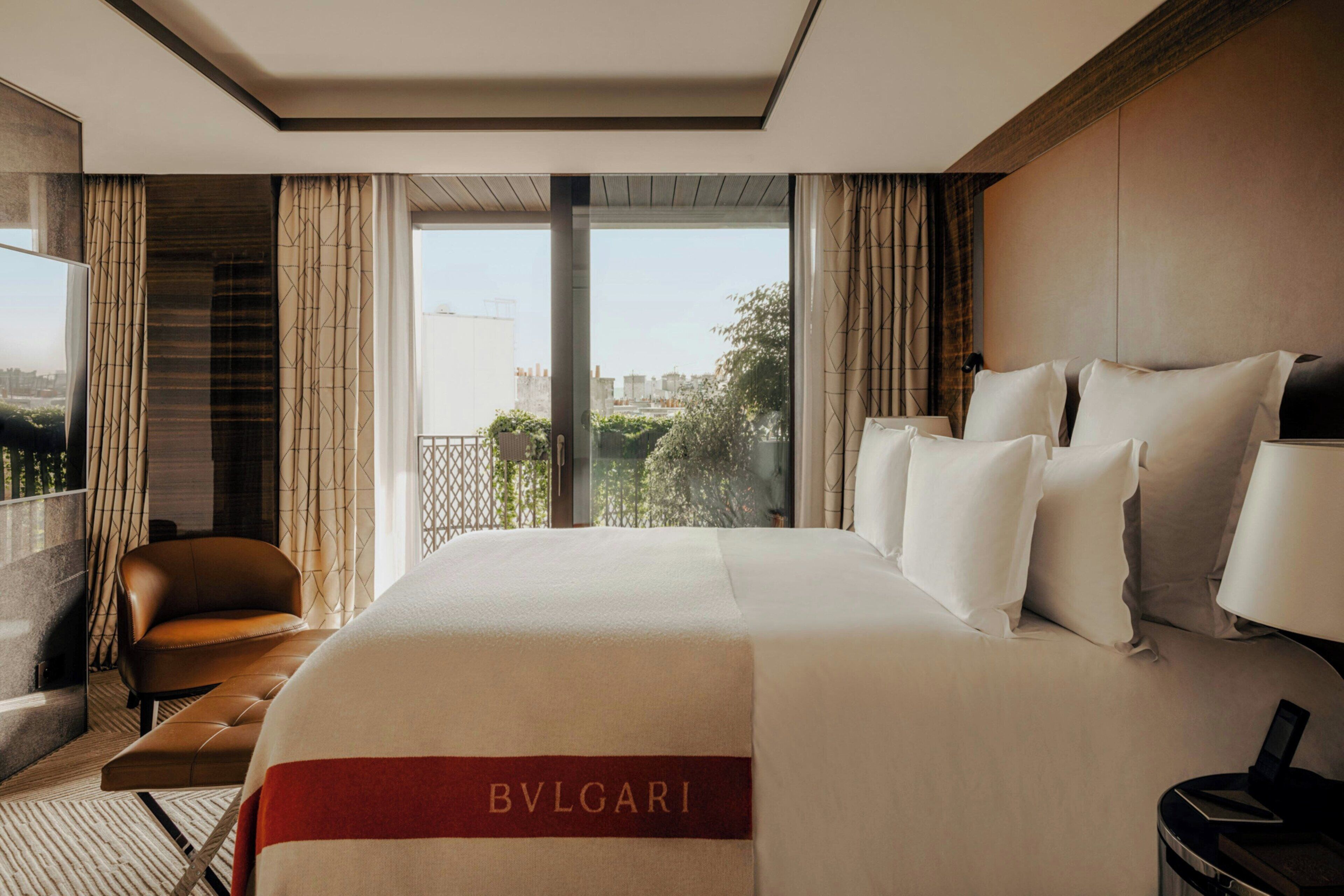 A hotel room with a large bed, a leather armchair, a small table, and a view of the city through large windows.