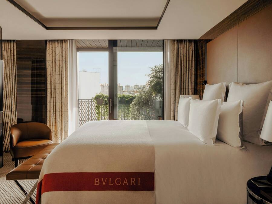 A hotel room with a large bed, a leather armchair, a small table, and a view of the city through large windows.