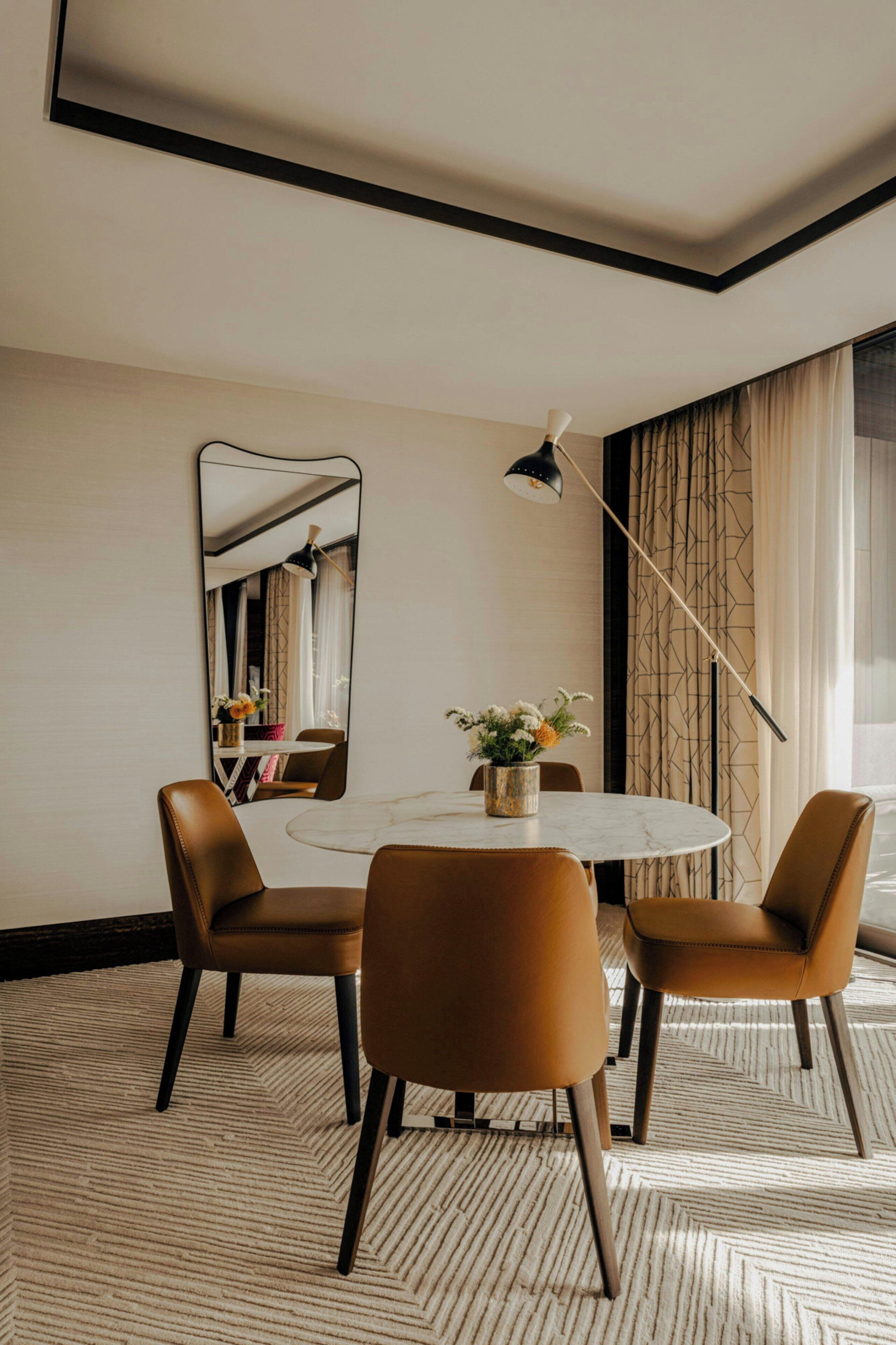 A dining area with a round table, two chairs, a mirror, a vase with flowers, and a lamp.