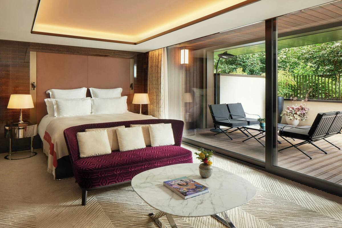 A modern hotel room with a large bed, a sofa, a small table, and a balcony with outdoor seating.