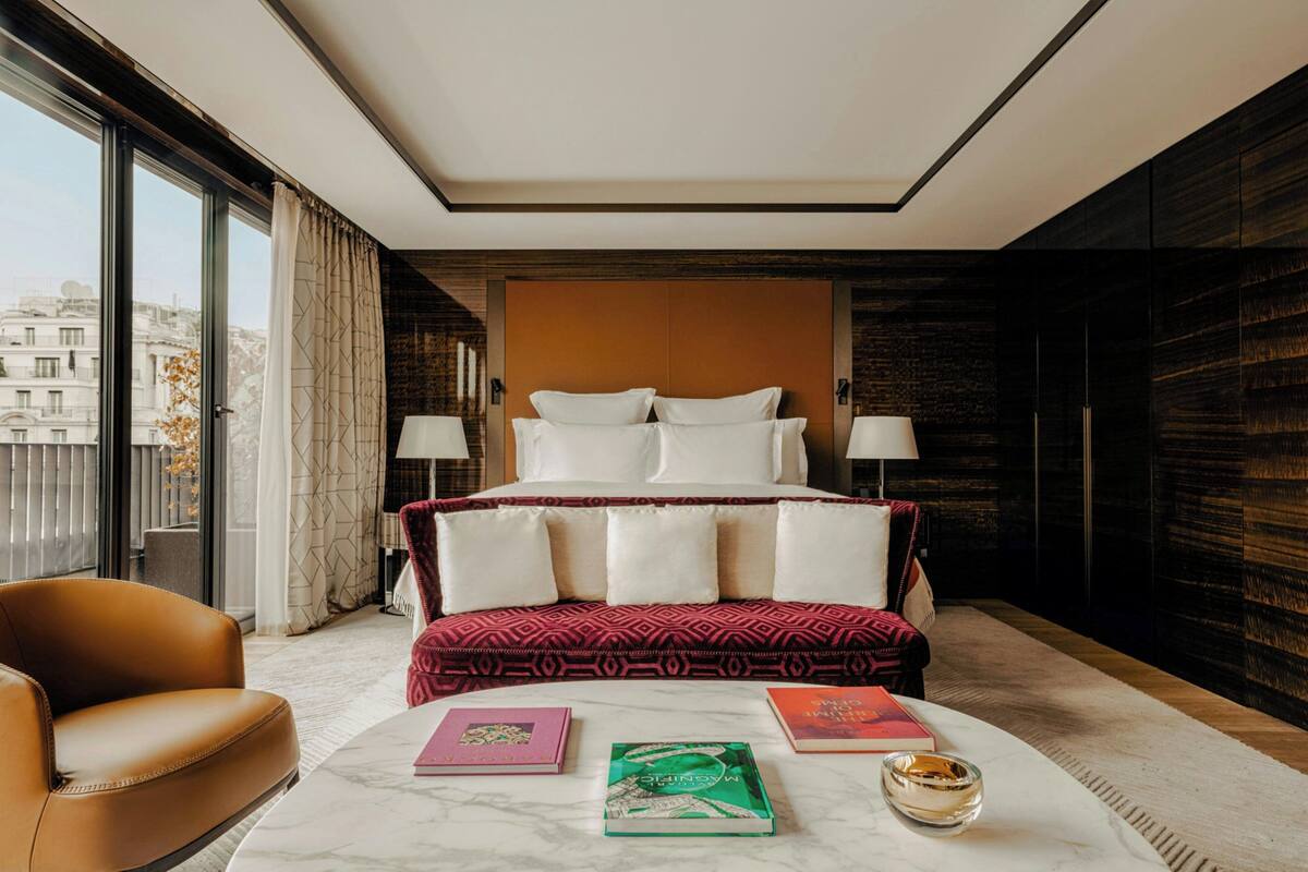 A modern hotel room with a large bed, two bedside lamps, a marble coffee table with books and a glass, and a view of the city through large windows.
