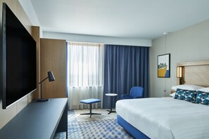 Habitación de hotel con cama, escritorio con televisor, silla, mesita pequeña y ventana con cortinas.