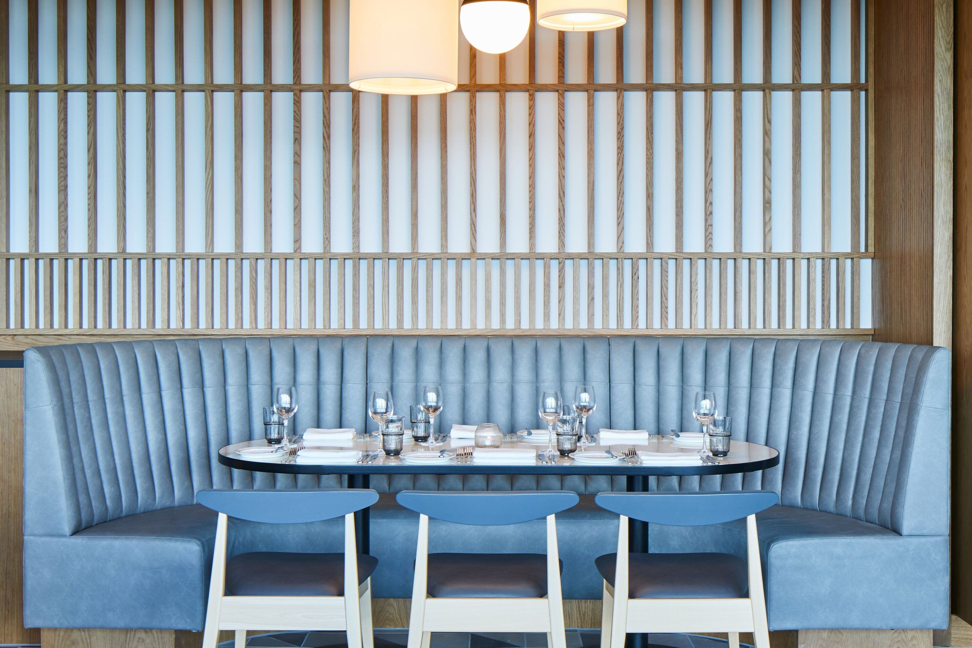 En restaurant med et blåt polstret båsarrangement, et rundt bord dækket med glas og tallerkener, og en væg med trælameller med hængende lys.