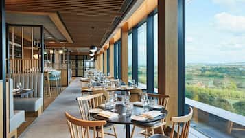 A restaurant with a long table and chairs, large windows, and a view of the countryside.