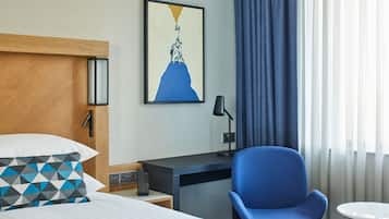 A hotel room with a bed, a desk, a chair, a small table, a lamp, and a painting on the wall.