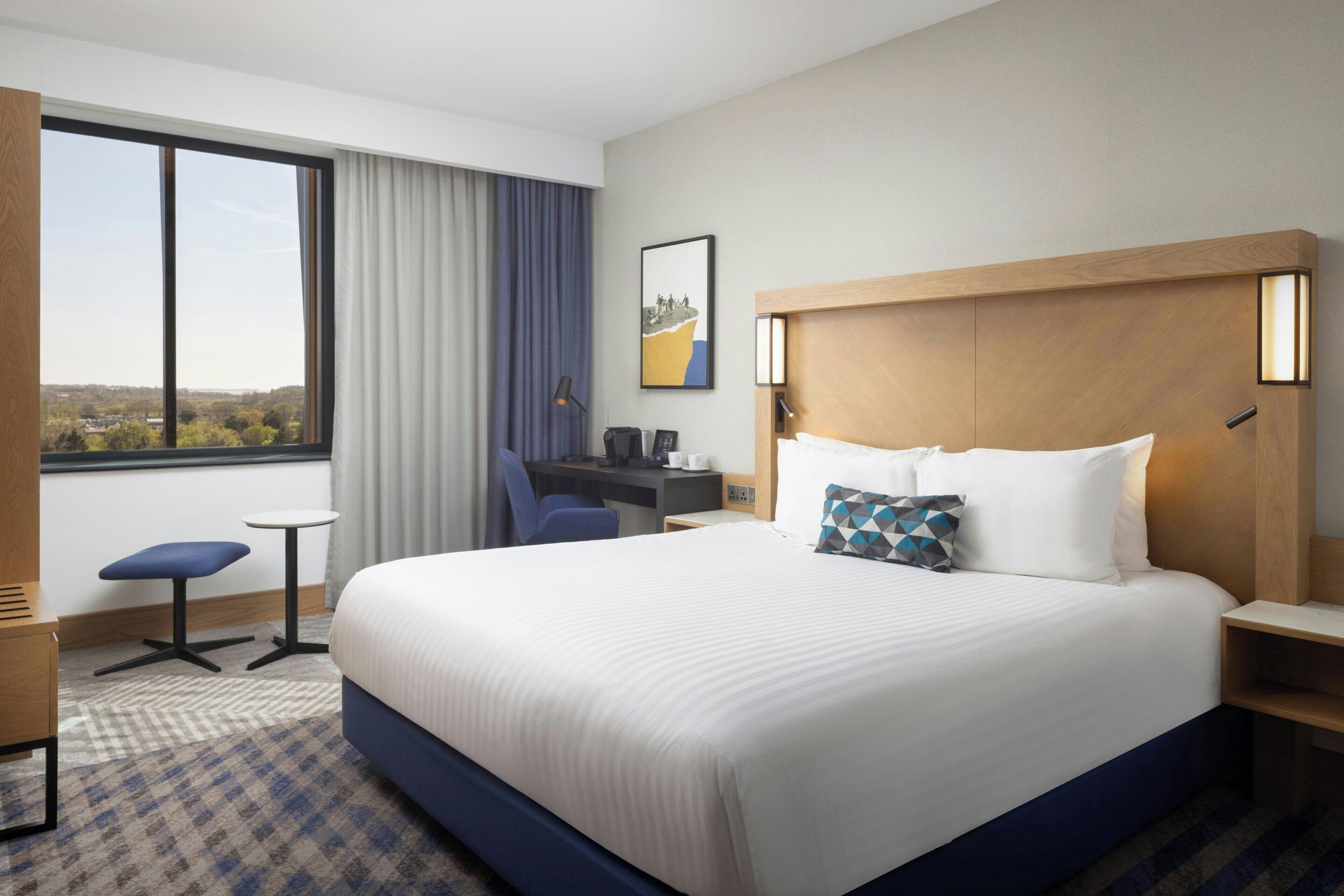 Ein Hotelzimmer mit einem großen Bett, einem Schreibtisch, einem Sessel, einem kleinen Tisch und einem fenster mit Vorhängen.