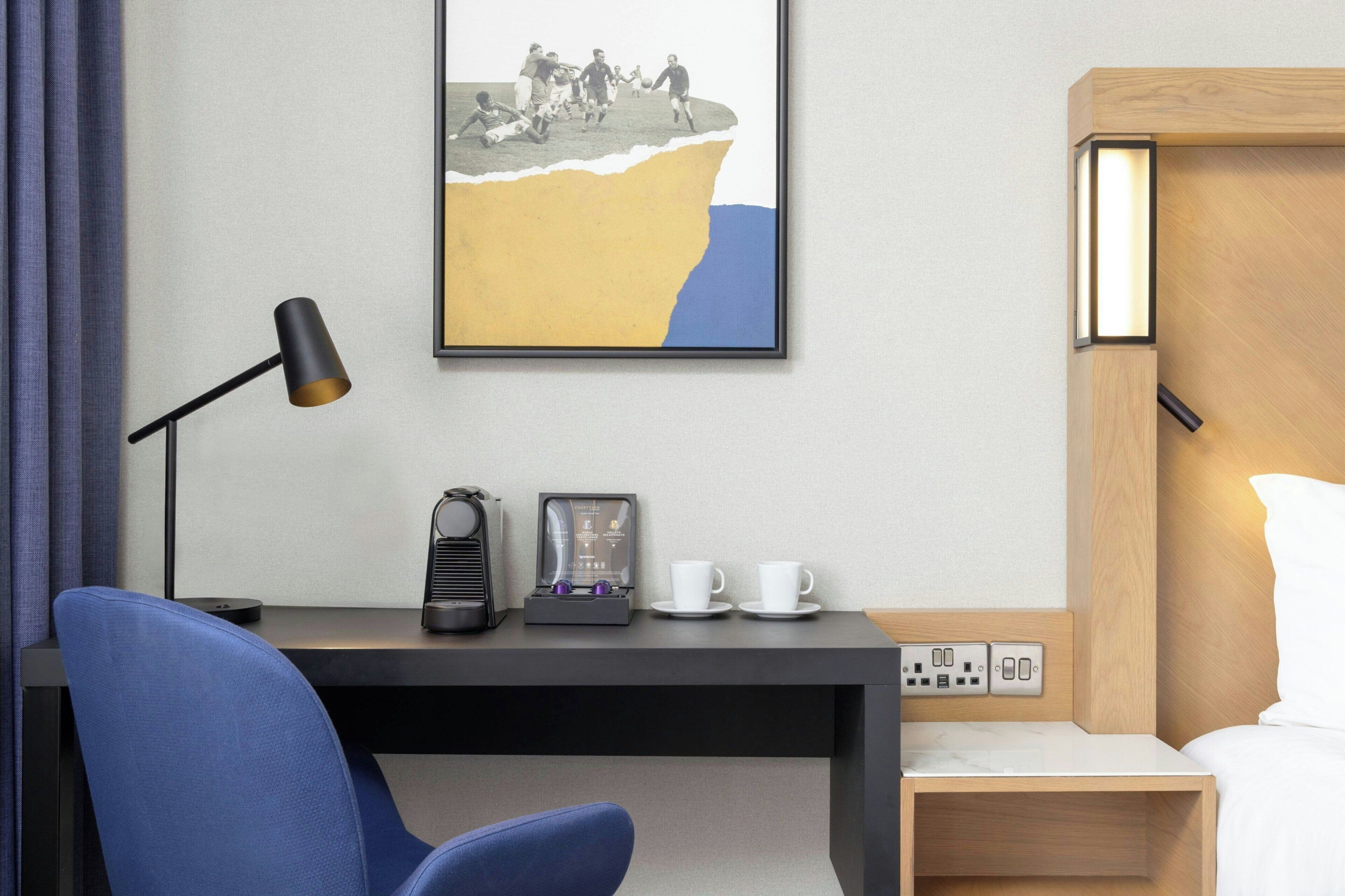 A hotel room with a desk, a chair, a bed, a bedside table with a lamp, a coffee maker, and a framed picture on the wall.