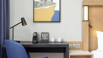 A hotel room with a desk, a chair, a bed, a bedside table with a lamp, a coffee maker, and a framed picture on the wall.