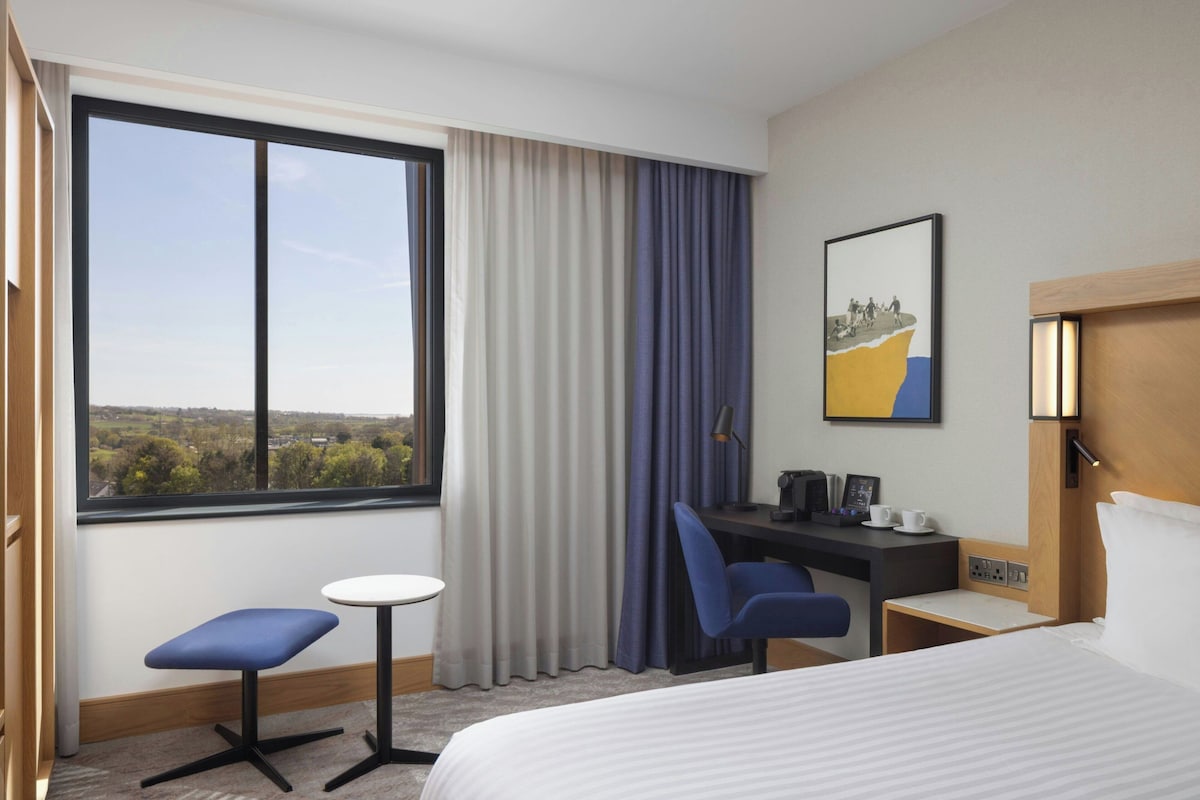 Habitación de hotel con cama, escritorio, silla, mesita, ventana con cortinas y un cuadro en la pared.