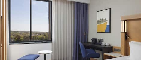 Ein Hotelzimmer mit Bett, Schreibtisch, Stuhl, kleinem Tisch, einem Fenster mit Vorhängen und einem Bild an der Wand.