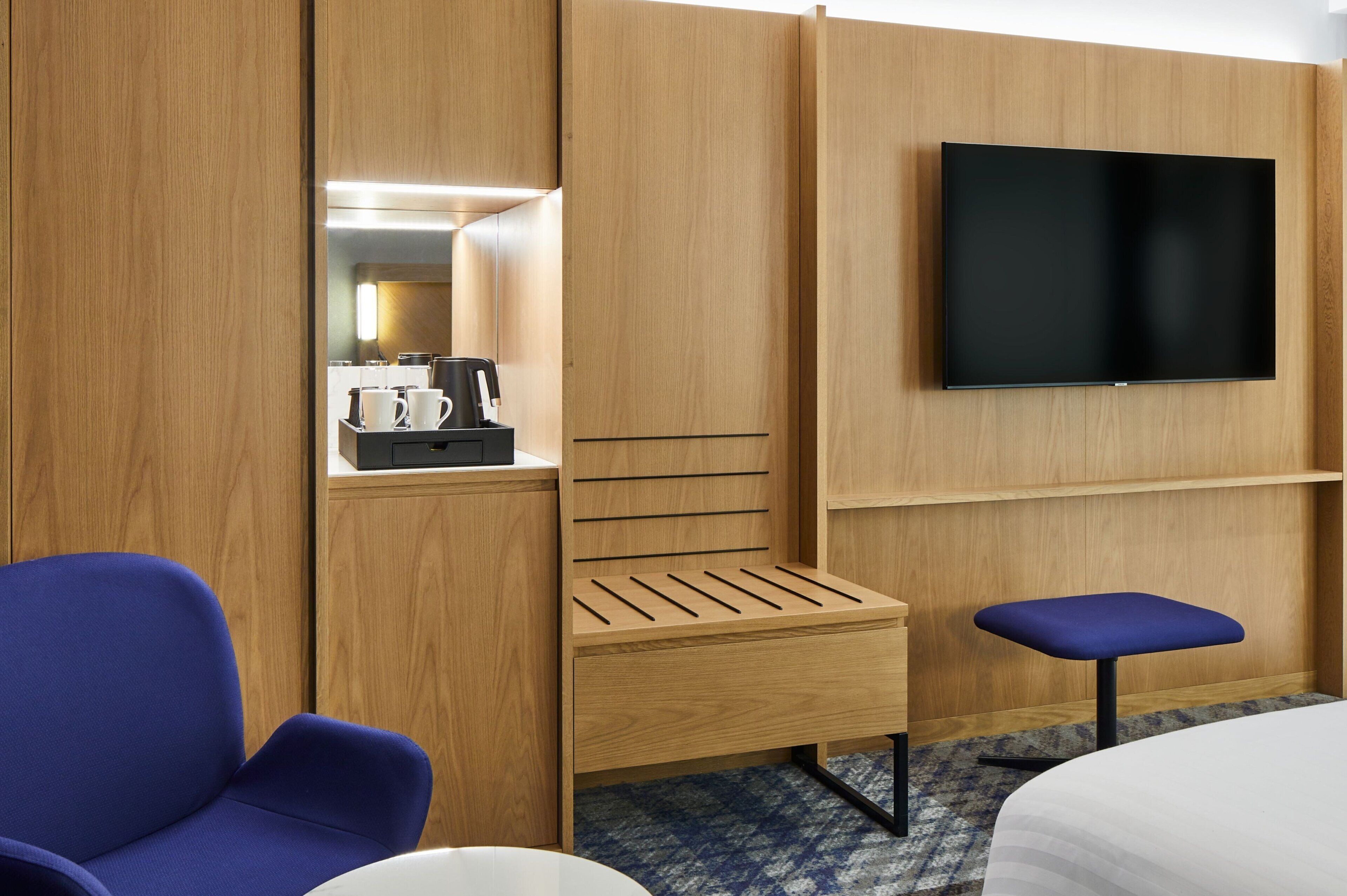 Habitación de hotel con armario de madera, televisión de pantalla plana, una silla azul, un banco de madera pequeño y una cafetera en el estante del armario.