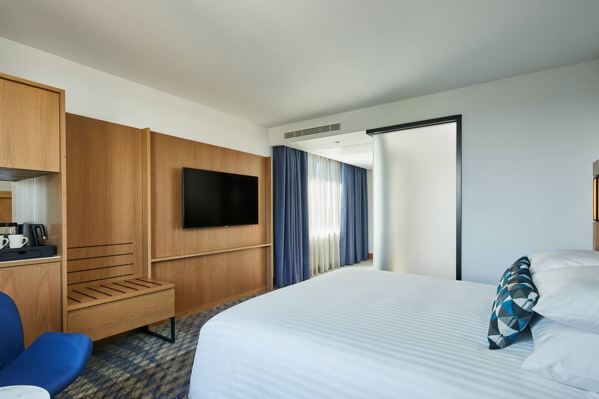 Habitació d'hotel amb un llit gran, televisió penjada a la paret, armari de fusta, zona de seients petita i porta al bany.